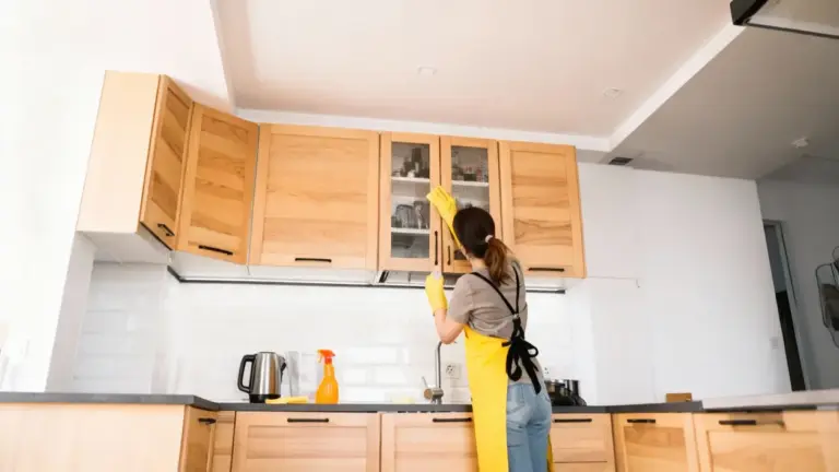 Persona che pulisce la parte superiore dei pensili della cucina con guanti e grembiule